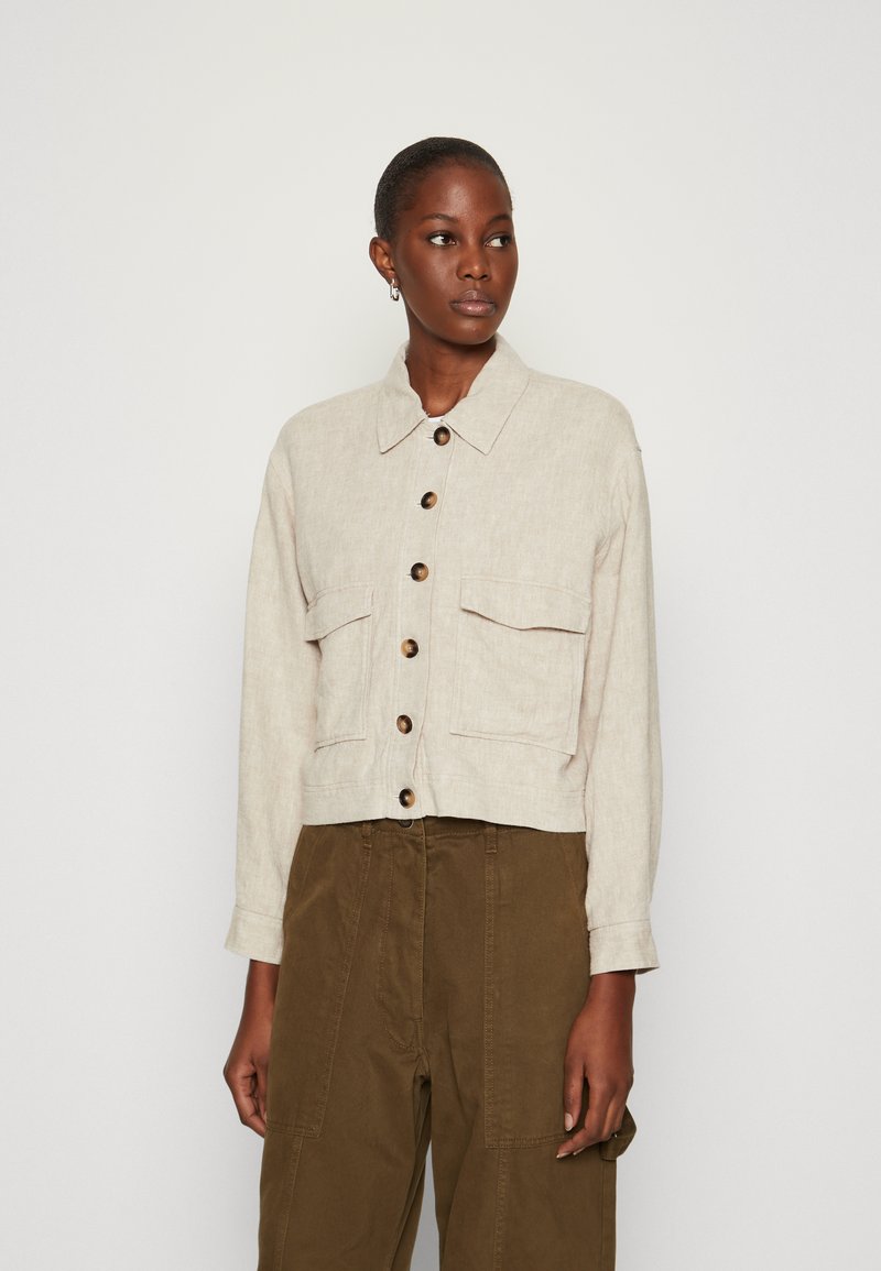 Beige linne cropped jack met een kraag, sluiting met knopen aan de voorkant, twee borstzakken en lange mouwen; gecombineerd met olijfgroene broeken.