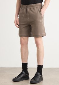 Shorts en coton marron avec une taille élastique, poches latérales et un patch logo. Associés à des baskets noires et des chaussettes côtelées.