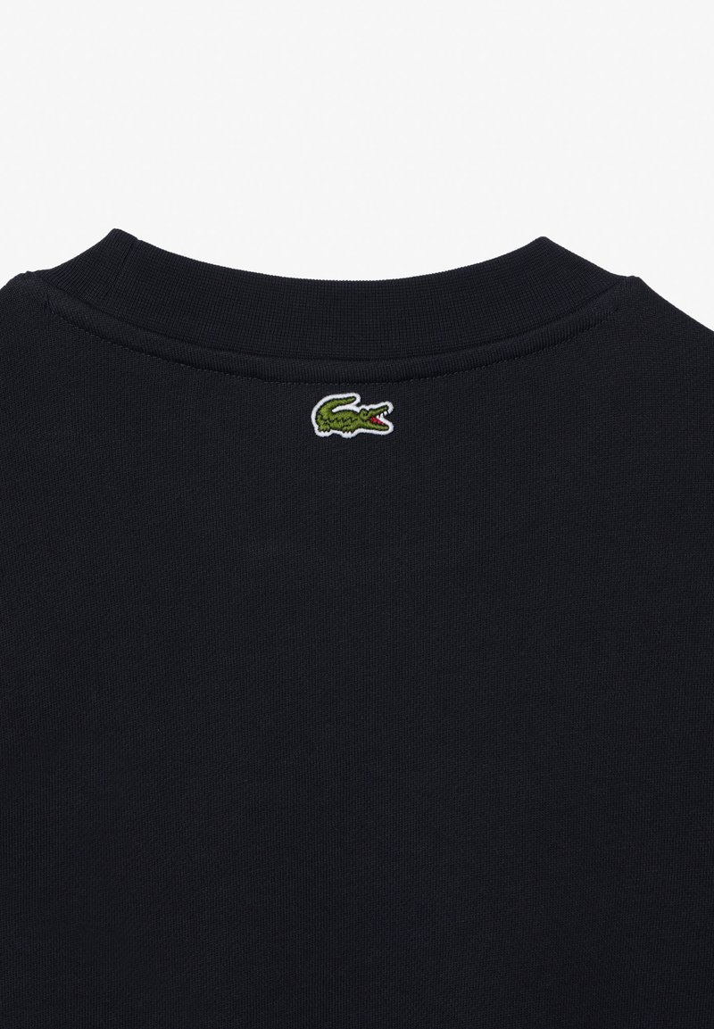 Sudadera de algodón negra con cuello acanalado y un pequeño logo de cocodrilo verde bordado en la parte posterior, cerca del cuello.