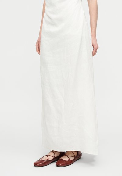 Λευκό μακρύ φόρεμα από λινό με άνετη γραμμή και διακριτή υφή, συνδυασμένο με μπορντό δερμάτινα φλατ παπούτσια με λουριά που σταυρώνονται.