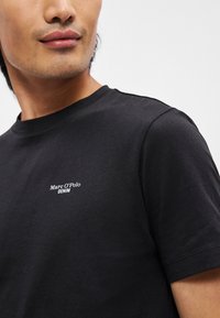 Melns kokvilnas T-krekls ar īsām piedurknēm, kurā ir balts teksta logo "Marc O'Polo DENIM" kreisajā krūšu daļā. Gluda tekstūra.
