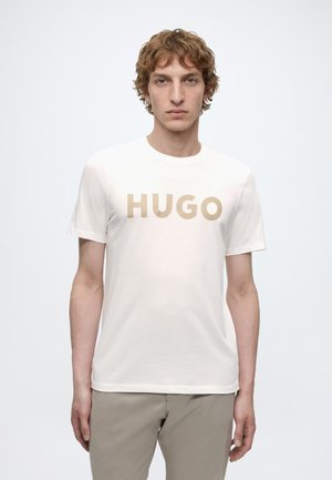 Giovane uomo con capelli ricci castano chiaro che indossa una maglietta bianca con la scritta beige "HUGO" e pantaloni beige, in piedi davanti a uno sfondo chiaro e uniforme.