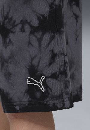 Shorts tie-dye noirs et gris avec un logo Puma blanc brodé près de l'ourlet, portés par une personne avec la peau des jambes visible.