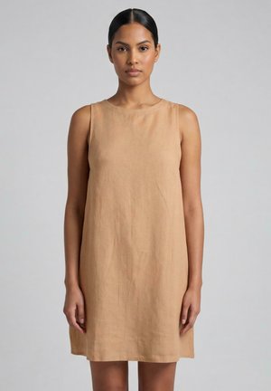 Donna con capelli scuri raccolti, che indossa un vestito beige senza maniche fino al ginocchio su uno sfondo grigio uniforme.
