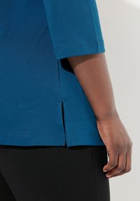 Top blu tealo con maniche corte, fessure laterali e una texture liscia. Mostrato di lato su uno sfondo chiaro, abbinato a pantaloni neri.