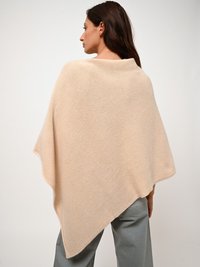 Beiger Poncho aus weichem Stoff, mit asymmetrischer Form und drapierten Kanten, getragen über grauen Hosen.