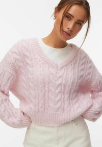 Jeune femme portant un pull en maille torsadée rose clair par-dessus un haut blanc, posant avec les mains sur les hanches devant un fond blanc.