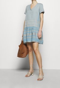 Světle modré šaty s krátkými rukávy a výstřihem do V, s tiered lemem a texturovanými vzory. Doplněné o hnědou tkanou tašku a sandály na řemínky.