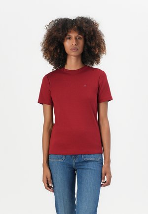 T-shirt rossa a maniche corte con un design aderente, realizzata in materiale morbido. Presenta un piccolo accento con marchio sul petto, abbinato a jeans blu.