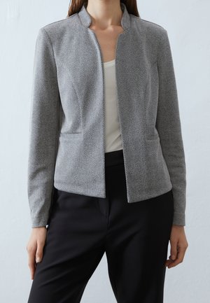 Vrouw draagt een grijze visgraatblazer over een witte top en zwarte broek tegen een effen lichte achtergrond.