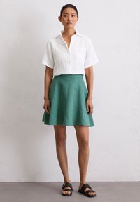 Witte linnen shirt met korte mouwen en een borstzak gecombineerd met een groene uitlopende rok. Zwarte instapsandalen maken de outfit compleet.