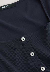 Gros plan sur un tissu côtelé foncé avec trois boutons blancs et une étiquette noire avec un texte blanc « "NEXT" » au niveau du col.