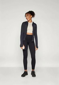 Svart idrottsjacka med långa ärmar, beige crop top och svarta leggings. Modellen bär svarta sneakers och håller en vattenflaska.