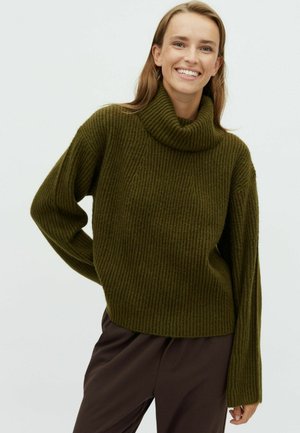 Hrubý rolákový svetr se žebrovaným vzorem v tmavě zelené barvě, vyrobený z texturované pleteniny. Má volný střih a široké rukávy. Kombinován s hnědými kalhotami.