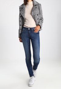 Abrigo de cuadros en blanco y negro con cuello con muesca sobre un suéter de punto beige, combinado con jeans ajustados oscuros y zapatillas blancas.