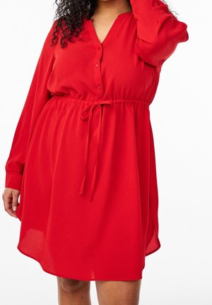 Femme portant une robe rouge à manches longues avec des boutons et une taille nouée, posant avec une main levée près de sa tête.
