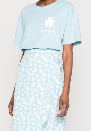 Henkilö pukeutuneena vaaleansiniseen paitaan, jossa on valkoinen päivänkakkara ja teksti "Life is good", yhdistettynä vastaavan kuosin omaavaan päivänkakkarahameeseen.