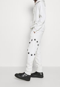 Grå mjukisbyxor med ett cirkulärt mönster av svarta stjärnor på sidan, kombinerade med en matchande grå hoodie och svarta sneakers.