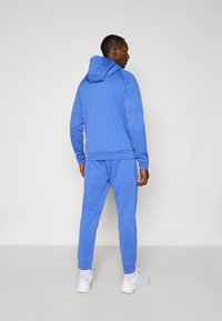 Blue cotton sweatshirt with hood and matching sweatpants. Features ribbed cuffs and a relaxed fit. Worn with white athletic shoes.