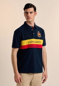 Polo navy de manga corta con cuello de dos botones, con una franja horizontal amarilla y roja, logotipo bordado y el número 2.