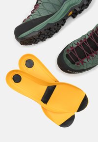 Traction-focused hiking shoe in dark green with maroon accents, paired with yellow insoles featuring black non-slip pads.