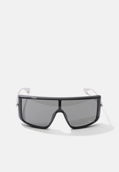 Saulesbrilles ar lielu, taisnstūra formu. Melna plastmasas rāmja ar tumši pelēkiem stikliem un baltām akcentētām kājām. Elegants dizains.