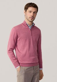 Pullover in rosa chiaro, realizzato in materiale morbido, presenta un colletto con zip, polsini e orlo a coste, indossato sopra una camicia a righe blu.