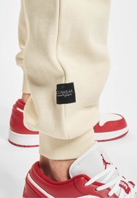 Pantalon de survêtement beige avec étiquette noire « Jocawear » au-dessus du poignet côtelé, porté avec des baskets Air Jordan blanches et rouges.
