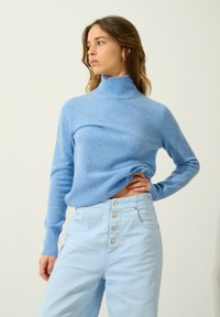 Hellblauer Rollkragenpullover aus weichem Strickstoff, taillierte Passform, mit langen Ärmeln. Kombiniert mit hellblauen Hochbundhosen mit Knopfdetails.