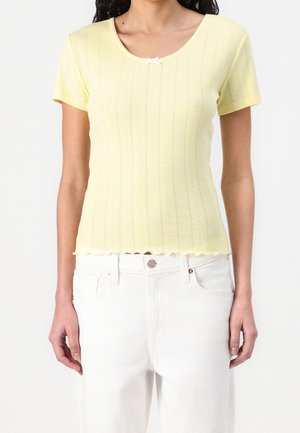 Femme portant un haut moulant à manches courtes côtelé jaune pâle avec un petit nœud blanc au niveau du col et un pantalon blanc.