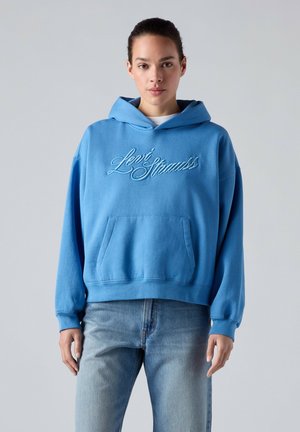 Junge Frau trägt einen blauen Levi Strauss Hoodie und blaue Jeans, steht vor einem schlichten hellgrauen Hintergrund.