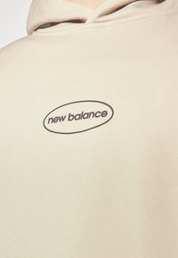 Felpa beige realizzata in tessuto morbido, con scollatura rotonda, cappuccio regolabile con coulisse, e un logo "new balance" nero all'interno di un ovale.
