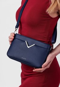 Námořnická kabelka z umělé kůže se stříbrným V-špice akcentem a nastavitelným popruhem, obsahující zipové uzavření a logo detail na přední straně.