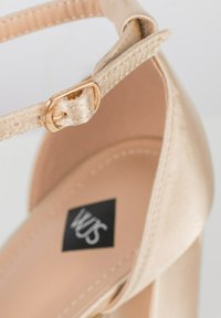 Beige High Heels Sandale mit satinierter Oberfläche, ausgestattet mit einem dünnen Knöchelriemen und einer goldfarbenen Schnalle. Schwarz-weißes Logo im Inneren.