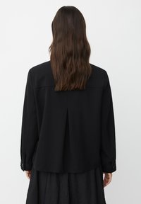 Persoon met lang bruin haar draagt een zwart shirt met lange mouwen en een gestructureerde zwarte rok, met de rug naar de camera gekeerd tegen een effen witte achtergrond.