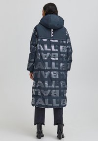 Abrigo acolchado azul marino con un prominente logo "BALL" en gris claro. Cuenta con capucha, mangas largas y longitud hasta debajo de la rodilla. Se ven vaqueros negros y botas.