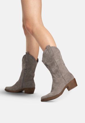 Light gray suede mid-calf cowboy boots with embroidered details and wooden block heels worn on crossed legs against white background.