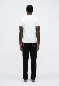 Vīrietis stāv ar muguru, ģērbies baltā T-kreklā, melnās biksēs ar izbalējušām aizmugurējām kabatām un melnbaltiem sporta apaviem uz baltas fona.