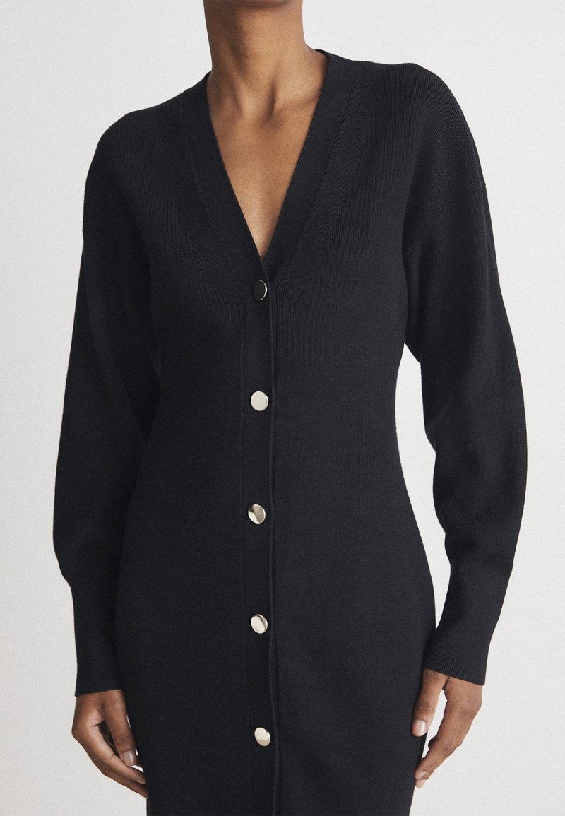 Robe en maille noire à manches longues avec encolure en V et boutons ronds argentés sur le devant, portée par une personne debout sur un fond uni.