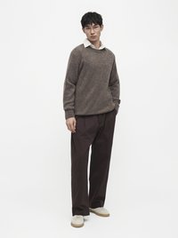 Homme portant un pull en maille beige sur une chemise blanche à col, un pantalon large marron foncé, des baskets beiges et des lunettes transparentes, debout devant un fond blanc.