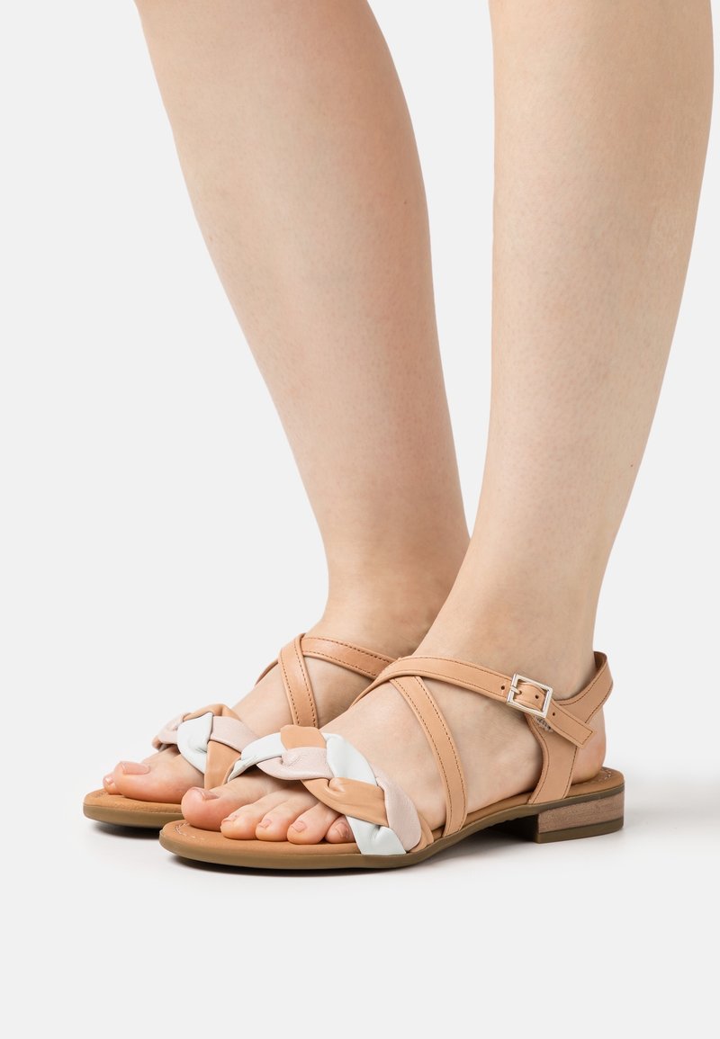 Pieds portant des sandales plates couleur beige avec des brides tressées blanches et rose clair et une fermeture à boucle à la cheville, sur un fond clair uni.