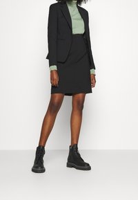 Donna che indossa un blazer nero e una gonna, un maglione a dolcevita verde e stivaletti platform neri con lacci, in piedi contro uno sfondo bianco.