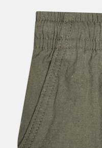 Tissu vert olive avec une finition texturée, doté d'une taille froncée et de détails de couture contrastants. Comprend une poche latérale.