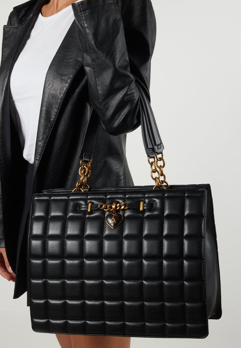 Sort quiltet håndtaske lavet af læder, prydet med guldfarvede detaljer, kædeaccenter og en hjerteformet charme. Inkluderer dobbelte skulderremme.