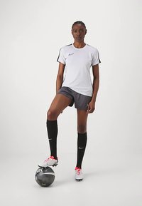 Nike'i sportlik riietus, mis sisaldab valget lühikeste varrukatega särki mustade detailidega, halli lühikest pükst, musti põlvikuid ja valgeid jalgpallikrampe, seisab musta palliga.