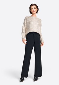 Helles beiges Strickpullover mit lockerer Passform und Rundhalsausschnitt, kombiniert mit weiten schwarzen Hosen und spitzen schwarzen Absätzen.