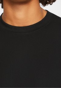 Dorosły w gładkiej czarnej koszulce z okrągłym dekoltem, pokazujący szyję i górną część klatki piersiowej, z widocznymi krótkimi kręconymi włosami.