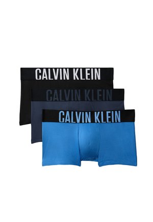 Junger Mann steht barfuß in dunkelgrauen Calvin Klein Boxershorts vor einfachem grauem Hintergrund.