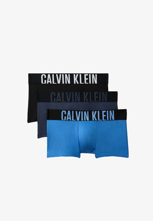 Młody mężczyzna stoi boso w ciemnoszarych bokserkach Calvin Klein na jednolitym szarym tle.