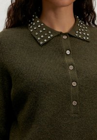Close-up van een olijfgroen gebreid sweater met een knoopsluiting en een kraag versierd met twee rijen grote heldere strassstenen aan elke kant.